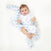 A baby smiles on a white background, wrapped in an aden + anais essentials cotton muslin swaddle blanket featuring blue paisley designs. Wearing a white outfit and looking up, the baby enjoys comfort and style from these breathable cotton essentials.
