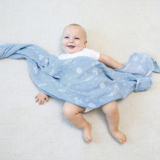A cheerful baby in a white onesie lies on a soft, light surface, partially covered with an aden + anais essentials cotton muslin swaddle blanket from the 4 pack, featuring breathable blue cotton muslin with white patterns.