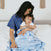 A woman with long dark hair sits on a bed, gently holding her baby swaddled in an aden + anais essentials cotton muslin swaddle blanket. Wearing a blue dress, she smiles at the little one in a white shirt. The breathable fabric and cozy room create an ambiance of warmth and love.
