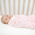 A baby is nestled in an aden + anais essentials cotton muslin swaddle blanket, featuring a pink and white pattern. With wide eyes and turned slightly to the side, the baby lies on a white surface with wooden bars in the background, offering a serene setting ideal for these swaddle essentials.