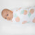 A baby lies on a white surface, wrapped snugly in an aden + anais Essentials Cotton Muslin Swaddle Blanket featuring a pineapple pattern in pink and green hues, gazing up with a calm expression.