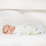 A baby wrapped in an aden + anais essentials cotton muslin swaddle blanket from the 4 pack with pastel floral patterns lies on a white bed, looking at the camera.