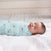A baby sleeps peacefully, wrapped snugly in an aden + anais essentials cotton muslin swaddle blanket, featuring small boat patterns. The baby lies on a soft, white surface in a bright room with a light wooden background.