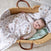 A baby smiles, lying in a wicker basket on a soft surface. Wrapped in an aden + anais essentials cotton muslin swaddle blanket with colorful patterns, the baby wears a light-colored outfit. Another patterned swaddle from the 2-pack is also inside the basket.