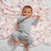 A baby, content with one hand in their mouth, lies on a colorful Disney™ essentials cotton muslin swaddle blanket by aden + anais. Crafted from soft, breathable fabric featuring cartoon characters, this cozy blanket adds playful designs to the scene.