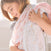 A young girl smiles, wrapped in an aden + anais essentials cotton muslin swaddle blanket. Her brown hair frames her content face as she relaxes snugly in the sunlit room.