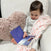 A toddler sits in a white chair, holding a blue-covered book. Theyre wrapped in an aden + anais essentials cotton muslin swaddle blanket from the 4 pack, nestled beside a fluffy heart-shaped pillow.