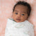 A baby with curly dark hair lies peacefully on a soft, peach-patterned blanket, wrapped in an aden + anais essentials cotton muslin swaddle featuring a breathable white fabric with a bunny print.