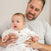 A man in light clothing holds a baby on his lap, both surrounded by aden + anais Disney™ essentials cotton muslin swaddle blankets. The baby, snug in breathable cotton muslin, gazes to the side while the man smiles warmly at them in a softly lit indoor setting.