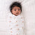 A dark-haired baby is snugly wrapped in a Disney™ essentials cotton muslin swaddle blanket by aden + anais, featuring colorful animal prints. The breathable cotton keeps the baby cozy on a textured white surface as they gaze upward with wide eyes.