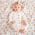 A baby in a cream-colored knitted onesie with wooden buttons is on a patterned blanket with small colorful figures. Wearing a matching headband, the infant looks up curiously, nestled among aden + anais Disney™ essentials cotton muslin swaddle blankets.