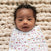 A baby in an aden + anais essentials Easy Swaddle Wrap Quick Change with adjustable fasteners, red stars, and yellow lightning patterns lies on a textured beige surface. The curly-haired baby calmly gazes at the camera.