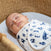 A peaceful baby sleeps in a crib, wrapped in an aden + anais essentials velboa wrap swaddle from the Quick Change 3 Pack, TOG 1.5. The swaddle features dark blue animal prints, and the crib has a natural woven texture; the babys expression is calm with eyes closed.