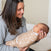A woman with long hair smiles at a baby cradled in her arms, wrapped in an aden + anais essentials easy swaddle adorned with moons and suns. The wrap, designed to prevent startle reflexes, pairs well with her cozy gray sweater as they enjoy their time indoors.