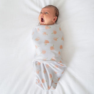 A baby rests on a white surface, wrapped in an aden + anais essentials easy swaddle with peach patterns. The baby gazes upward, mouth open in surprise or mid-yawn. The swaddles adjustable fasteners allow for arms-in or arms-out use, while the babys short dark hair adds to the pure innocence.