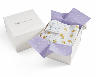 A white Gift Box by HALO, partially open with purple tissue paper, reveals a folded white baby blanket featuring blue trim and a lemon print.