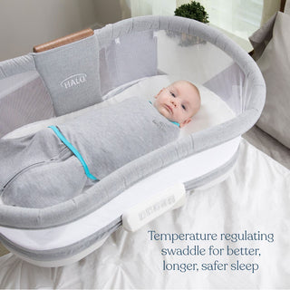A baby rests in a HALO Swaddle Ideal Temp TOG 1.5, snuggled in gray with IntelliThread for temperature regulation, atop white sheets in a mesh-sided bassinet. Text reads: Swaddle Ideal Temp TOG 1.5 for better, longer, safer sleep.