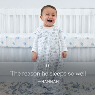 A smiling baby stands in a crib wearing the HALO Eric Carle The Hungry Caterpillar 100% Cotton Wearable Blanket TOG 0.5. White crib with blue-patterned bedding in the background. Text: The reason he sleeps so well — Hannah.