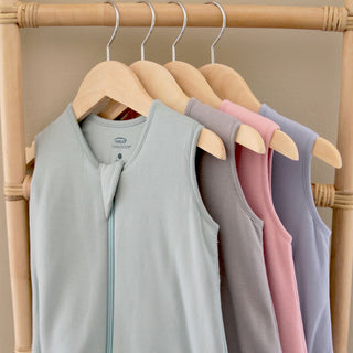 Four HALO SuperSoft Bamboo Wearable Blanket TOG 1.5 sleep sacks in pastel colors (light blue, beige, pink, and lavender) hang on wooden hangers on a bamboo rack, each crafted with a buttery 3-layer bamboo design for comfort.
