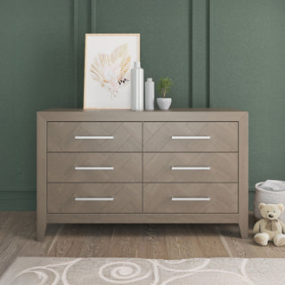 The Child Craft Kieran 3 Piece Nursery Set, featuring a light wood dresser with six drawers and sleek silver handles, is displayed against a green wall for a modern touch that complements any nursery. The dresser is styled with a framed abstract artwork, two vases, and a small plant on top, while a teddy bear rests on the floor beside it and a soft rug lies in the foreground.