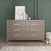 The Child Craft Kieran 3 Piece Nursery Set, featuring a light wood dresser with six drawers and sleek silver handles, is displayed against a green wall for a modern touch that complements any nursery. The dresser is styled with a framed abstract artwork, two vases, and a small plant on top, while a teddy bear rests on the floor beside it and a soft rug lies in the foreground.