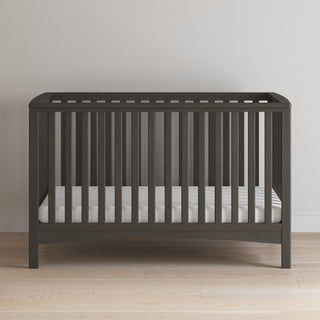 A dark brown Child Craft London 2-Piece Nursery Set crib with vertical slats stands against a light wall on a light wood floor, featuring a white mattress inside.