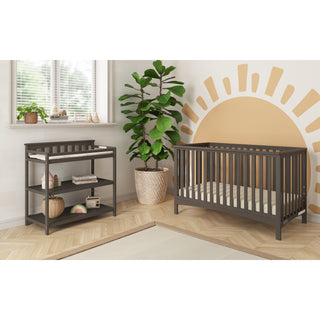 A modern nursery with the Child Craft London 2-Piece Nursery Set—crib and changing table—plus a leafy plant in a woven pot, sun mural by the blinded window, light wood floors, and cozy decor for a stylish space.