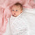 A baby with wide blue eyes is cozily wrapped in an aden + anais essentials cotton muslin swaddle blanket, highlighting its breathable fabric. Dressed in a white blanket with grey and pink dots, the child rests on a white surface, partially covered by a soft pink textured blanket.