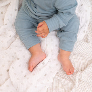 A baby in light blue ribbed pajamas sits on a textured white blanket with gray polka dots. Only the babys legs, feet, and a hand are visible, while an aden + anais essentials cotton muslin swaddle blanket from the 2 pack is gently draped nearby.