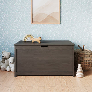 A Child Craft Scout 4 Piece Nursery Set—including a crib, dresser, glider, and toy box—features a dark wood toy box that sits in a nursery with toys, teddy bears, a basket, and wall art on light wood flooring and blue wallpaper with white branches, creating a cozy and coordinated space that complements the rest of the nursery furniture.