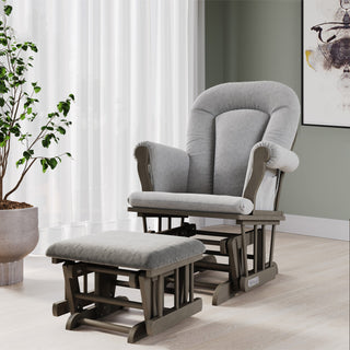 A cozy nursery featuring the Child Craft Hampton Arch Top 3 Piece Nursery Set—including a stylish gray upholstered glider chair with matching ottoman—sits on a light wood floor beside a potted plant and a window adorned with sheer white curtains. Abstract art accents the pale green wall, completing the serene space.