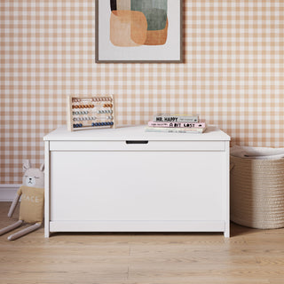 A white Child Craft Scout 4 Piece Nursery Set toy box sits in a room with gingham wallpaper, a framed abstract art piece above, books and an abacus on top, a plush cat toy, and a woven basket to the right—perfectly complementing the crib, dresser, and glider from this cozy and organized nursery furniture set.