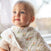 A light-haired child looks upward, wearing a soft pink outfit and an aden + anais essentials cotton muslin burpy bib. A floral-patterned blanket is draped over their shoulder against a softly blurred background, suggesting natural light through a window.