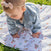 A baby in a denim jacket and a headband with a bow is crawling on the soft aden + anais essentials cotton muslin dream blanket, spread over the grass.