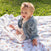 A smiling baby sits outdoors on an essentials cotton muslin dream blanket by aden + anais, wearing a denim jacket, a light outfit, and a pink bow headband. The lush green grass background creates a charming and serene setting.