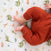 A baby in a rust-colored onesie is snuggled on the Breathable Nursery Bundle by HALO, lying on a white blanket adorned with green, orange, and blue dinosaur patterns.