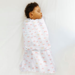 A baby peacefully sleeps in a HALO Swaddle 100% Cotton TOG 1.5, nestled in a white blanket with pink and orange rainbows. The adjustable wings provide comfort and promote healthy hip development, covering the baby entirely except for the head against a soft white surface.