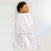 A baby peacefully sleeps in a HALO Swaddle 100% Cotton TOG 1.5, nestled in a white blanket with pink and orange rainbows. The adjustable wings provide comfort and promote healthy hip development, covering the baby entirely except for the head against a soft white surface.
