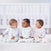 Three babies are seated on a soft white carpet in front of a crib, each wrapped in cozy HALO Wearable Blanket Micro-Fleece TOG 1.0. One sports a whale pattern, another has stripes, and the third shows small orange designs. These blankets offer safe sleep as the little ones curiously gaze at each other.
