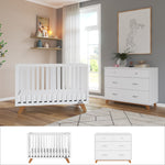 A contemporary nursery showcases the Child Craft Soho 2-Piece Nursery Set with Crib and Dresser, both in white with wooden legs, complemented by neutral walls, stylish shelving, greenery, framed artwork, and a large window that fills the space with natural light.