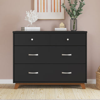 A sleek black dresser with four drawers, silver handles, and wooden legs stands against a pink wall, capturing the clean lines of mid-century modern design. Carefully arranged atop are decorative accents—a white coral sculpture, a vibrant plant, and framed artwork—offering the perfect finishing touches for your contemporary nursery. Pair this look seamlessly with the Child Craft Soho 3-Piece Nursery Set with Crib, Dresser and Chest to elevate your space or complement other pieces from the collection.