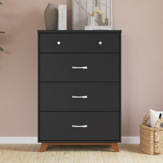 A black dresser from the Child Craft Soho 3-Piece Nursery Set with Crib, Dresser, and Chest stands against a light pink wall. Decorative items such as books, a vase, and artwork are arranged on top. To the right sits a woven basket filled with pillows, while a plant is partially visible on the left.