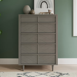 A gray dresser with five drawers from the Child Craft Surrey Hill 3-Piece Nursery Set (which includes a crib, dresser, and chest) stands against a green wall. On top of the dresser are a vase, decorative objects, and framed abstract art. A patterned rug is visible in the foreground, perfectly complementing other coordinating pieces from this elegant nursery furniture collection.