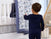 A toddler in navy pajamas stands by a white crib in the cozy nursery, reaching for a hanging quilt with blue animal motifs and muslin details. The aden + anais essentials cotton muslin bedding features a starry design, while a basket and shelves sit in the background.