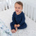 A smiling toddler in blue pajamas sits on a white crib mattress with a star pattern, nestled in the cozy nursery adorned with the aden + anais essentials cotton muslin bedding set 3 pack.
