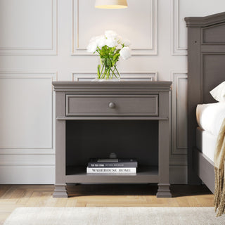 A Child Craft Westgate Nightstand in Chelsea Gray, featuring a drawer and open shelf with a book, sits by the bed. A glass vase of white flowers rests on top, beneath a hanging lamp. The room has light walls with molding and wooden floors.