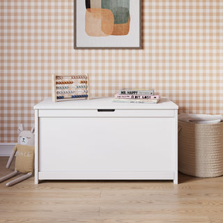 The Child Craft Harmony 33" Kids Toy Box in matte white sits against a brown gingham wallpapered wall, topped with an abacus, books, and framed art. On the wooden floor lie a plush bunny and woven basket, creating a charming, child-safe storage space.