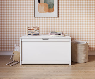 The Child Craft Harmony 33" Kids Toy Box in matte white sits against a brown gingham wallpapered wall, topped with an abacus, books, and framed art. On the wooden floor lie a plush bunny and woven basket, creating a charming, child-safe storage space.
